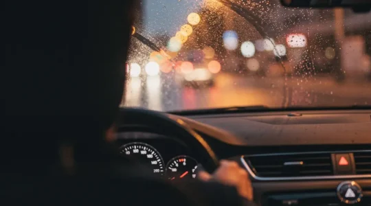 Vue cinématique depuis l'intérieur d'une voiture la nuit par temps de pluie, suggérant une atmosphère de conduite sous tension.
